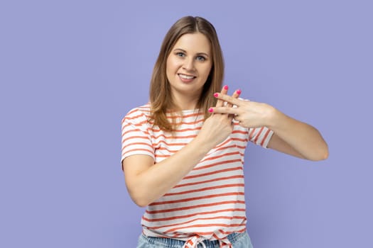 Portrait of blond woman wearing striped T-shirt making hashtag sign with fingers, tagging posts in social networks, recommending tags. Indoor studio shot isolated on purple background.