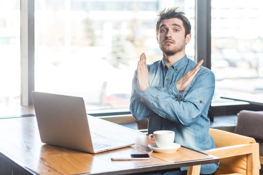 Portrait of serious handsome young man freelancer in blue jeans shirt working on laptop, showing x sign, no way gesture, looking at camera. Indoor shot near big window, cafe background.