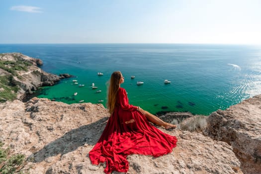 Woman red dress sea. Happy woman in a red dress and white bikini sitting on a rocky outcrop, gazing out at the sea with boats and yachts in the background