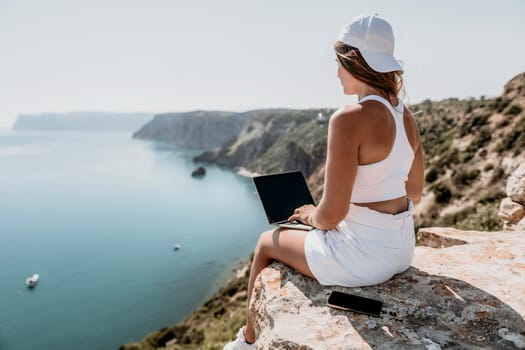 Digital nomad, Business woman working on laptop by the sea. Pretty lady typing on computer by the sea at sunset, makes a business transaction online from a distance. Freelance remote work on vacation