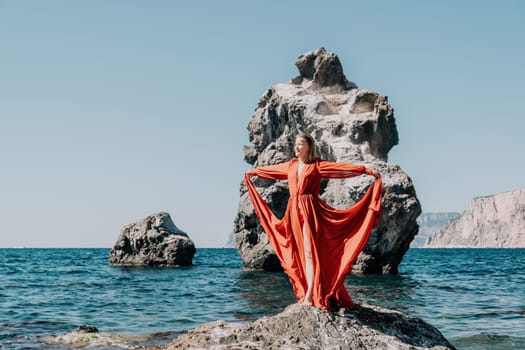 Woman travel sea. Young Happy woman in a long red dress posing on a beach near the sea on background of volcanic rocks, like in Iceland, sharing travel adventure journey