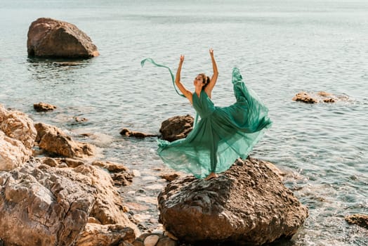 Woman green dress sea. Female dancer in a long mint dress posing on a beach with rocks on sunny day. Girl on the nature on blue sky background