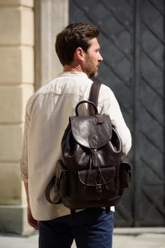 Handsome businessman walking on the street, with luxury leather backpack. Man wearing blue pants, sneakers, white t-shirt and beige shirt