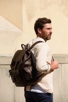 Handsome businessman walking on the street, with luxury leather backpack. Man wearing blue pants, sneakers, white t-shirt and beige shirt