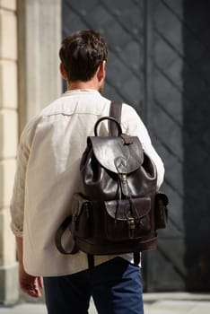 Handsome businessman walking on the street, with luxury leather backpack. Man wearing blue pants, sneakers, white t-shirt and beige shirt