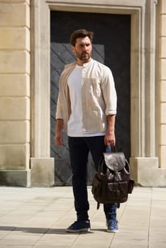 Handsome businessman walking on the street, with luxury leather backpack. Man wearing blue pants, sneakers, white t-shirt and beige shirt