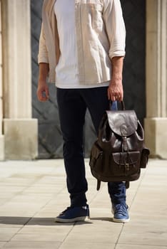 Handsome businessman walking on the street, with luxury leather backpack. Man wearing blue pants, sneakers, white t-shirt and beige shirt