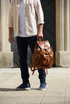 Handsome businessman walking on the street, with luxury leather backpack. Man wearing blue pants, sneakers, white t-shirt and beige shirt