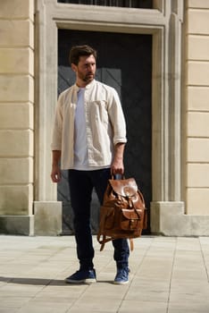 Handsome businessman walking on the street, with luxury leather backpack. Man wearing blue pants, sneakers, white t-shirt and beige shirt