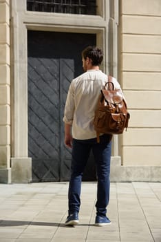 Handsome businessman walking on the street, with luxury leather backpack. Man wearing blue pants, sneakers, white t-shirt and beige shirt