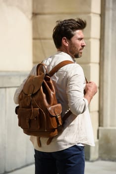 Handsome businessman walking on the street, with luxury leather backpack. Man wearing blue pants, sneakers, white t-shirt and beige shirt