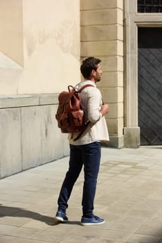 Handsome businessman walking on the street, with luxury leather backpack. Man wearing blue pants, sneakers, white t-shirt and beige shirt