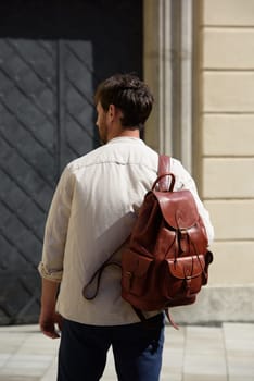 Handsome businessman walking on the street, with luxury leather backpack. Man wearing blue pants, sneakers, white t-shirt and beige shirt