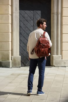 Handsome businessman walking on the street, with luxury leather backpack. Man wearing blue pants, sneakers, white t-shirt and beige shirt