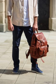 Handsome businessman walking on the street, with luxury leather backpack. Man wearing blue pants, sneakers, white t-shirt and beige shirt