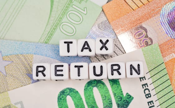 Small white beads with black letters TAX RETURN on euro banknotes, closeup detail from above.