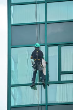 Industrial window cleaner - man hanging on roofs with safety equipment, cleaning facade of tall modern glass building.