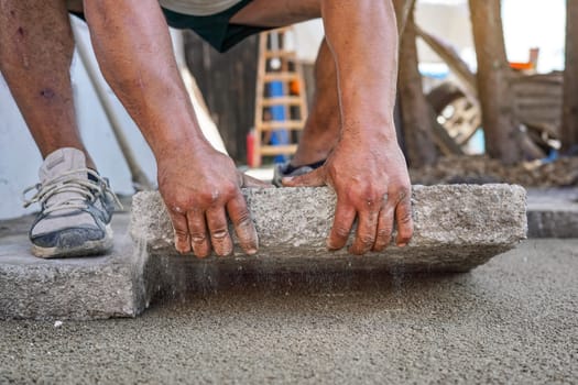 Installing new pavement or floor outside from large concrete tiles, closeup detail on male worker fitting stone block over sand and gravel base layer.