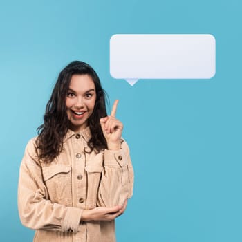 Cheerful millennial caucasian lady brunette student in casual showing finger up pointing at white blank communication balloon, got idea, isolated on blue background. Woman sharing her ideas