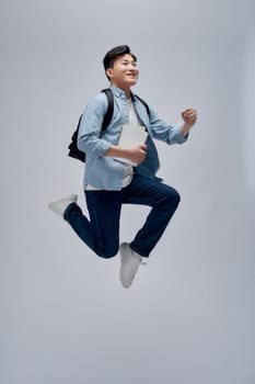 Full length portrait of a smiling young asian man carrying backpack isolated, holding books, jumping