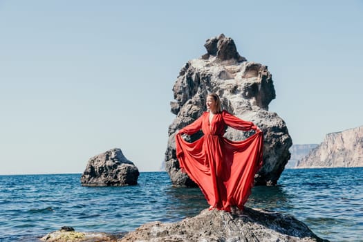 Woman travel sea. Young Happy woman in a long red dress posing on a beach near the sea on background of volcanic rocks, like in Iceland, sharing travel adventure journey