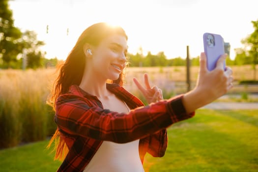 Red haired young blogger takes a selfie and shows peace at sunset using a smartphone