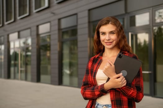 Happy red haired student, wearing casual clothes, holding a tablet, and standing near the university.