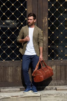 a man with a bag. black leather travel bag, street photo. man is wearing blue sneakers, jeans, a white T-shirt and a jacket.