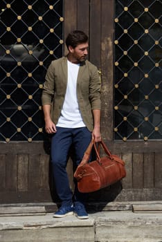 a man with a bag. black leather travel bag, street photo. man is wearing blue sneakers, jeans, a white T-shirt and a jacket.