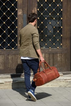 a man with a bag. black leather travel bag, street photo. man is wearing blue sneakers, jeans, a white T-shirt and a jacket.