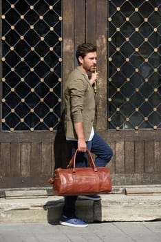 a man with a bag. black leather travel bag, street photo. man is wearing blue sneakers, jeans, a white T-shirt and a jacket.