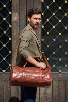 a man with a bag. black leather travel bag, street photo. man is wearing blue sneakers, jeans, a white T-shirt and a jacket.