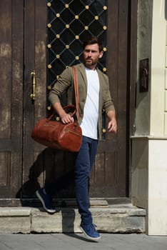 a man with a bag. black leather travel bag, street photo. man is wearing blue sneakers, jeans, a white T-shirt and a jacket.