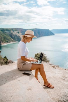 Freelance women sea working on the computer. Good looking middle aged woman typing on a laptop keyboard outdoors with a beautiful sea view. The concept of remote work