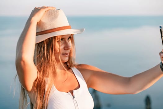 Selfie woman in hat, white tank top and shorts makes selfie shot mobile phone post photo social network outdoors on sea background beach people vacation lifestyle travel concept