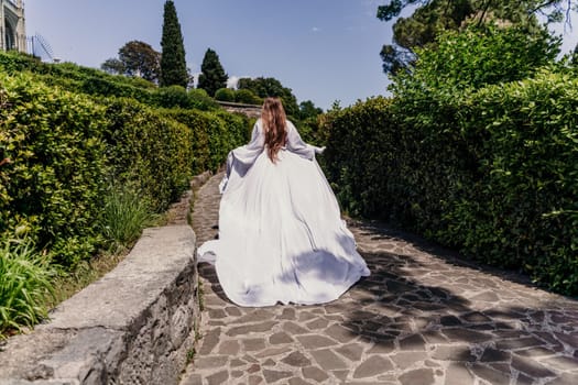 Brunette runs white dress park. A beautiful woman with long brown hair and a long white dress runs along the path along the beautiful bushes in the park, rear view.