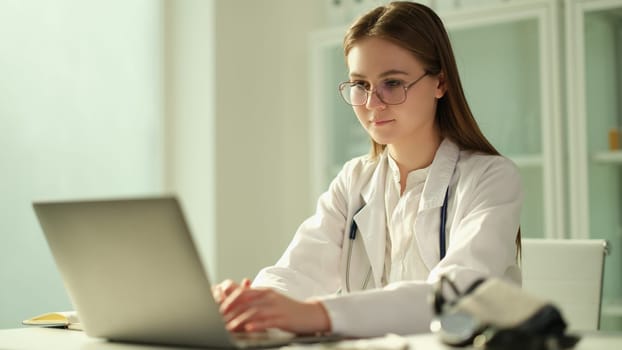 Focused female doctor sits at table in hospital and works on laptop online. Concentrated nurse or therapist looking at computer screen and consulting patient on gadget