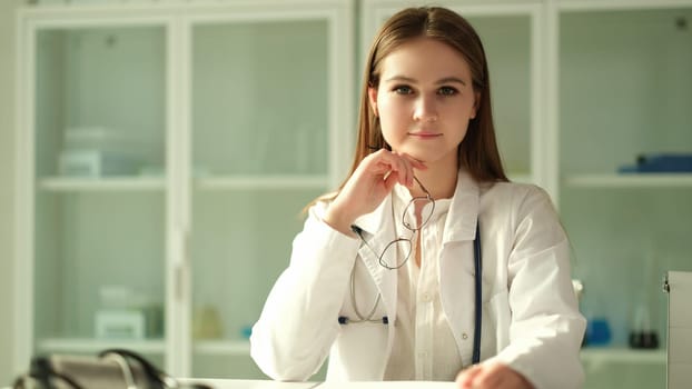 Portrait of smiling young female doctor at workplace holding eyeglasses in clinic. Nurse intern at hospital