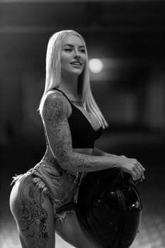 Sexy blonde woman model with silicone breasts and tattoos posing with motorcycle helmet in her hands, black and white photo