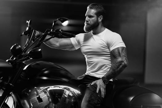 Sexy handsome attractive sporty muscular fitness model breaded biker in white tight tshirt sits on brutal beautiful motorcycle in the parking, black and white photo