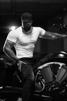 Sexy handsome attractive sporty muscular fitness model breaded biker in black cap and white tight tshirt sits on brutal beautiful motorcycle in the parking, black and white photo