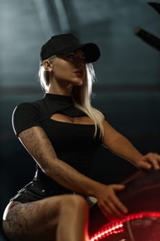 Sexy blonde woman with open neckline poses on motorcycle in the parking lot. Beautiful fitness model with tattoos sits on motorbike