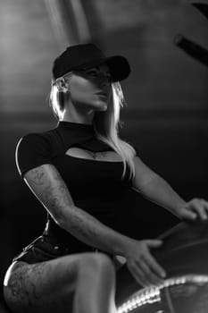 Sexy blonde woman with open neckline poses on motorcycle in the parking lot. Beautiful fitness model with tattoos sits on motorbike, black and white photo