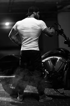 Sexy handsome attractive sporty muscular fitness model breaded biker with brutal beautiful motorcycle in the parking posing with a motorbike with his back to the frame, black and white photo