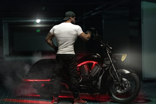 Sexy handsome attractive sporty muscular fitness model breaded biker with brutal beautiful motorcycle in the parking posing with motorbike with his back to the frame