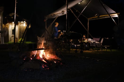 Bonfire near camping tent outdoors in evening and group of people sitting in background.