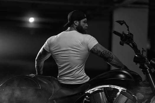 Sexy handsome attractive sporty muscular fitness model breaded biker with brutal beautiful motorcycle in the parking posing with a motorbike with his back to the frame, black and white photo