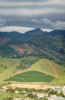 A mesmerizing aerial view of a green valley in the mountains with a small town surrounded by vast coffee plantations under a cloudy blue sky.