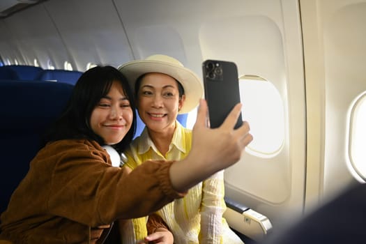 Smiling middle aged woman and daughter sitting in passenger airplane and taking picture, waiting for airplane landing.