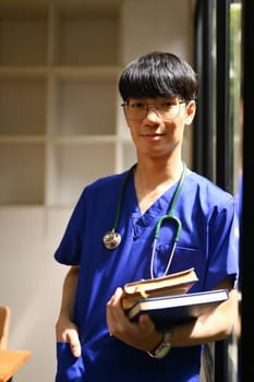 Portrait of medical student man wearing blue scrubs with stethoscope standing near window at campus.